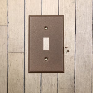 May include: A walnut brown metal light switch plate with a single light switch on a wood panel wall.