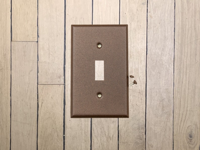 May include: A walnut brown metal light switch plate with a single light switch on a wood panel wall.