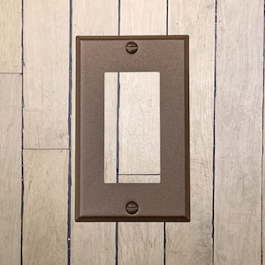 May include: A walnut brown metal wall plate with a rectangular opening for an electrical outlet. The plate has two small screws on the top and bottom.