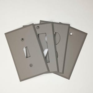 May include: A collection of four gray light switch covers. The covers have different cutouts for switches and outlets. The covers are made of a matte material and are arranged in a slightly overlapping pattern against a white background.