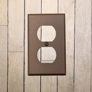 May include: A walnut brown metal electrical outlet cover plate with a single outlet cutout.
