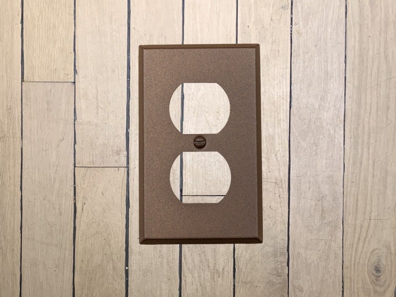 May include: A walnut brown metal electrical outlet cover plate with a single outlet cutout.
