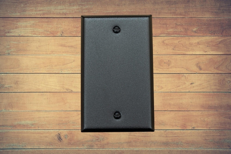 A single black metal blank switch plate with matching screws installed shown on a brown wood background.