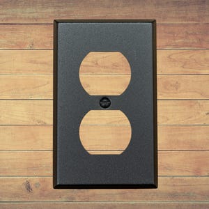 A single black metal duplex plate with two openings and a matching screw installed. It has a brown wood background.
