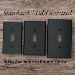 Three black metal toggle switch plates next to each other on a brown wood background. The words" Standard/Mid/Oversized" are shown above the plates, and "Also available in round corner" is written below the plates.