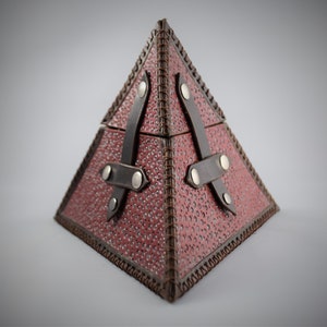 May include: A red leather pyramid-shaped box with brown leather straps and metal accents. The box has a stitched edge and a textured surface.