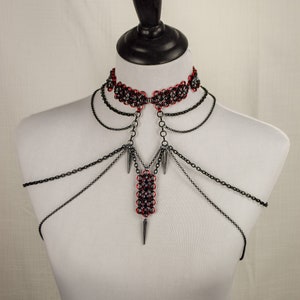 May include: A black chainmail choker necklace with a red and silver chainmail pendant. The pendant is attached to the choker with a silver chain. The necklace is on a white mannequin.