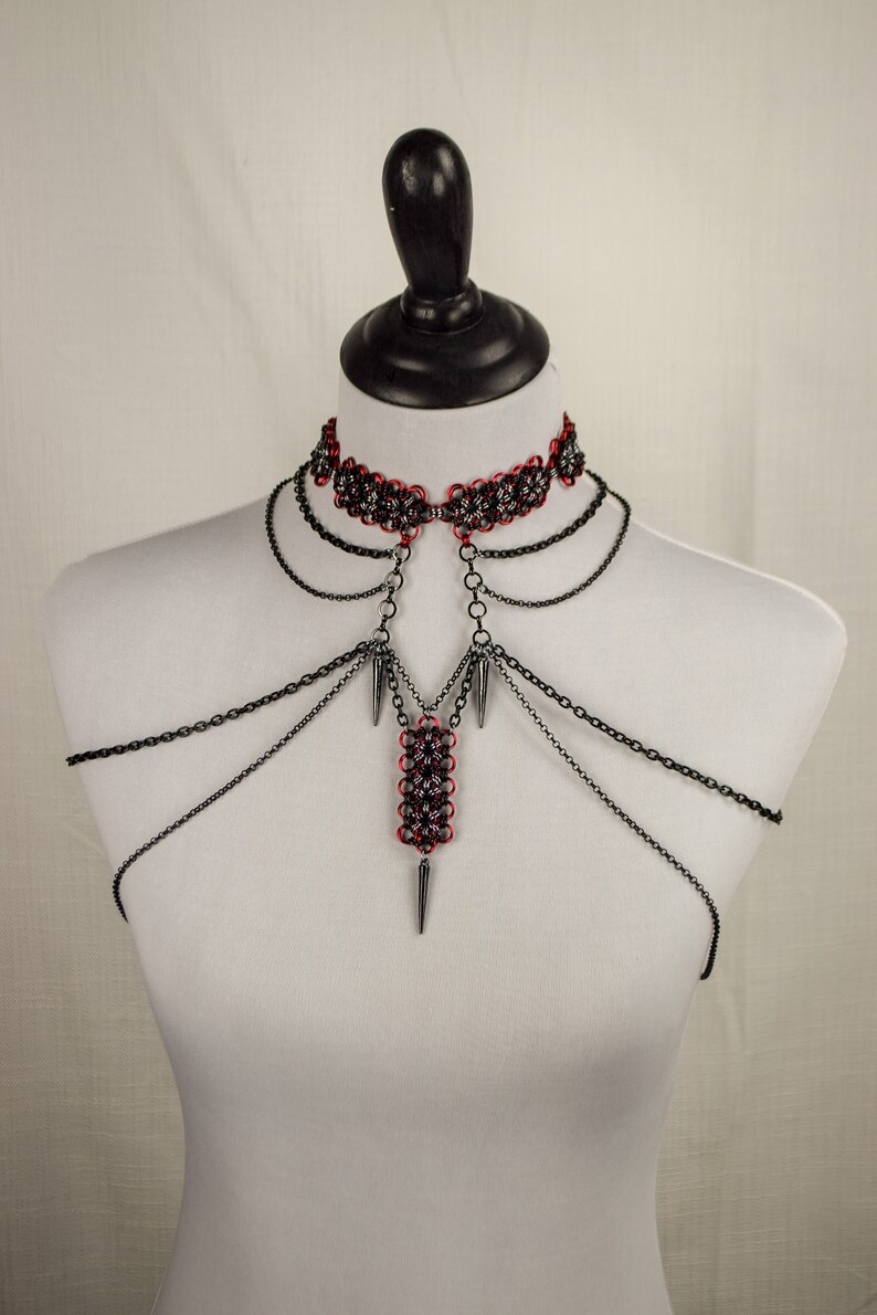 May include: A black chainmail choker necklace with a red and silver chainmail pendant. The pendant is attached to the choker with a silver chain. The necklace is on a white mannequin.
