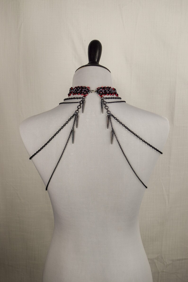 May include: A black chain choker necklace with red and black beads and silver spikes. The necklace is on a white mannequin.