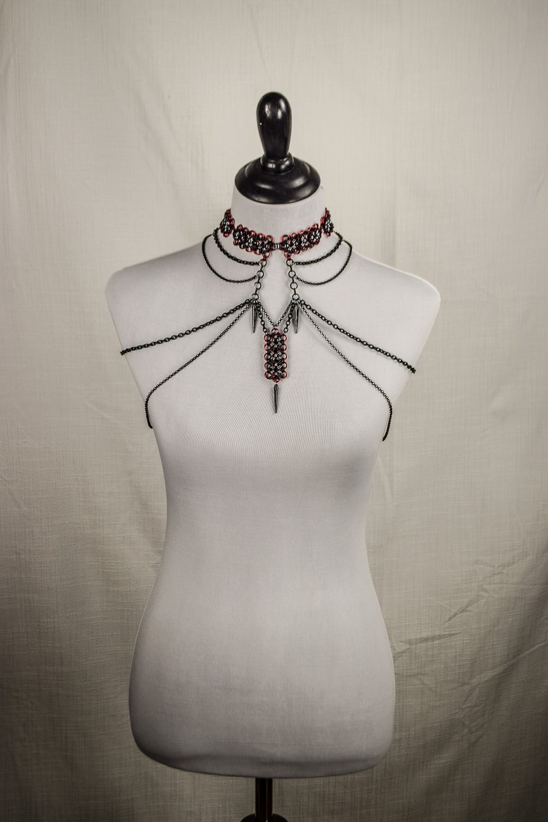 May include: A silver chain body harness with a red and black beaded choker. The harness has multiple chains that connect to a central pendant with a pointed tip.