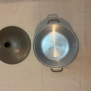 May include: An aluminum cooking pot with a lid. The pot is round with two handles and a lid with a black knob. The pot and lid are silver in color. The lid is to the left of the pot.