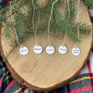 May include: Five silver necklaces with round pendants on a wooden surface. The pendants have the words "2020", "WORTHY", "D & B", "BRAVE", and "MAMA BEAR" engraved on them.