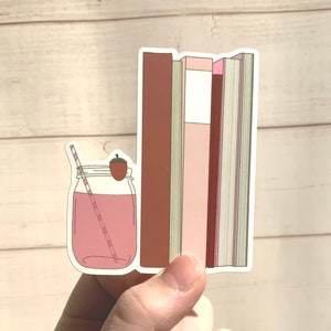 Puede incluir: Una pegatina que representa una bebida rosa en un tarro con una fresa y una pajita, junto a una pila de libros en tonos rosas y marrones. La pegatina se sostiene en una mano sobre un fondo de madera clara.