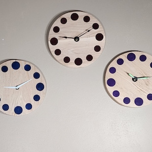 Puede incluir: Cuatro relojes de madera con puntos de colores diferentes alrededor de la esfera. Los relojes tienen agujas blancas y están colgados en una pared beige.