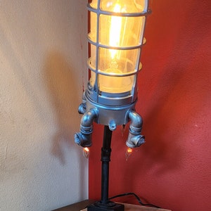 May include: A silver and black industrial style table lamp with a caged light bulb and three additional light bulbs on the base. The lamp is on a wooden base.