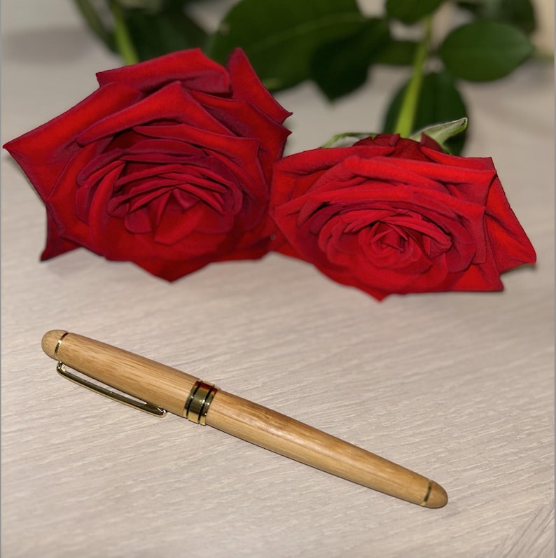 Puede incluir: Un bol&iacute;grafo de madera con detalles dorados descansa sobre una superficie clara, con dos rosas rojas en el fondo. El bol&iacute;grafo tiene un acabado de veta de madera natural y un clip dorado. Las rosas est&aacute;n en plena floraci&oacute;n.