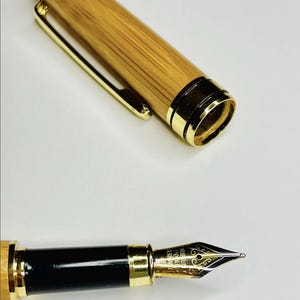 Puede incluir: Una pluma estilogr&aacute;fica con un cuerpo de bamb&uacute; y detalles dorados. La tapa de la pluma est&aacute; quitada, revelando el plum&iacute;n. El plum&iacute;n es dorado con dise&ntilde;os intrincados. La pluma tiene una secci&oacute;n de agarre negra.