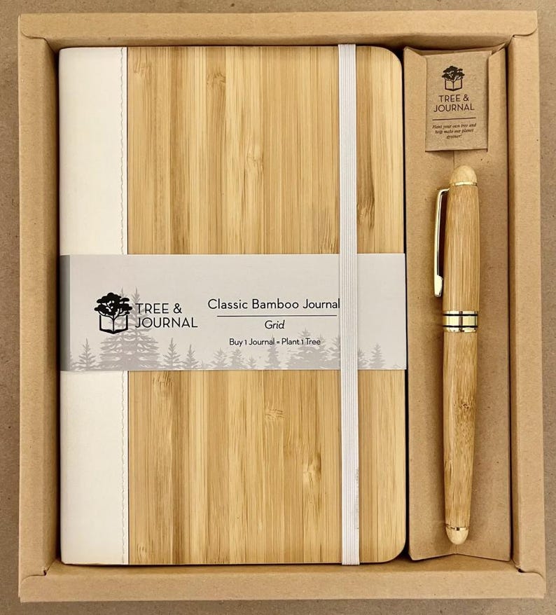 Puede incluir: Un juego de diario y bol&iacute;grafo de bamb&uacute; en una caja de cart&oacute;n. El diario es de color marr&oacute;n claro con una lomo y banda el&aacute;stica blancos. El bol&iacute;grafo tambi&eacute;n es de color bamb&uacute; con detalles dorados. El texto del diario dice "Classic Bamboo Journal".
