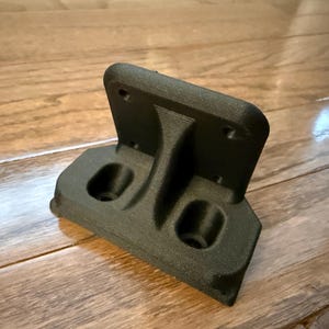 Op de afbeelding: Een zwarte, 3D-geprinte beugel met een getextureerd oppervlak. Het heeft een rechthoekige basis met twee cirkelvormige openingen en een verticale steun die leidt naar een bovenplaat met vier gaten. De beugel is ontworpen voor montage.