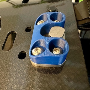 May include: A blue 3D printed part with four holes and a silver metal plate in the center. The part is attached to a black carbon fiber surface.
