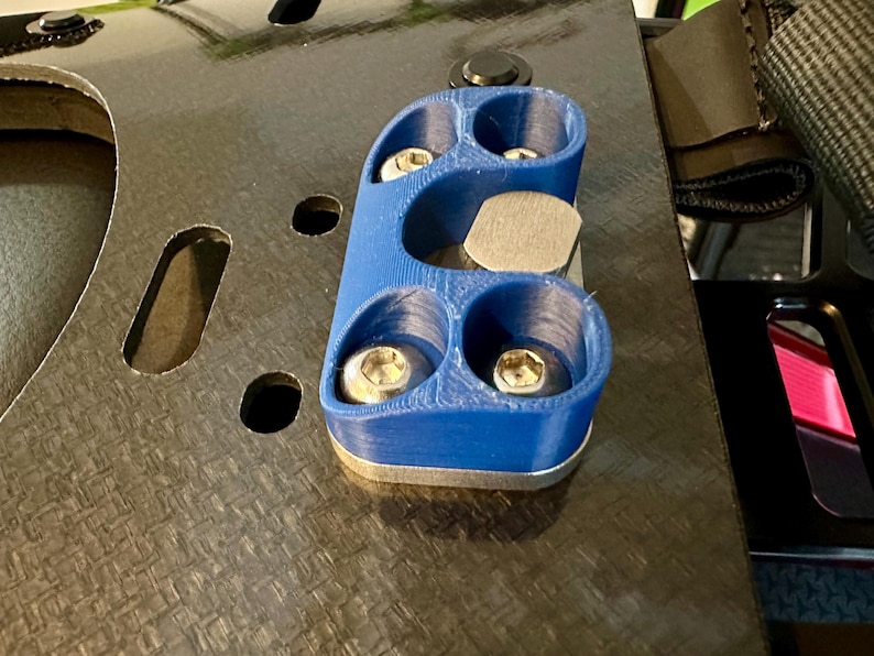 May include: A blue 3D printed part with four holes and a silver metal plate in the center. The part is attached to a black carbon fiber surface.