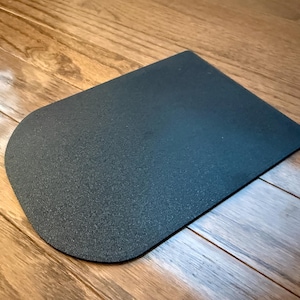 Könnte beinhalten: Eine dunkelgraue, rechteckige Matte mit abgerundeter Ecke. Die Matte hat eine strukturierte Oberfläche und liegt auf einem Holzboden.