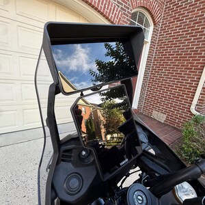 May include: A black motorcycle dashboard assembly featuring a large, rectangular upper display screen with a protective sun visor and a secondary, smaller vertical display screen below it, both showing reflections of the surrounding environment.