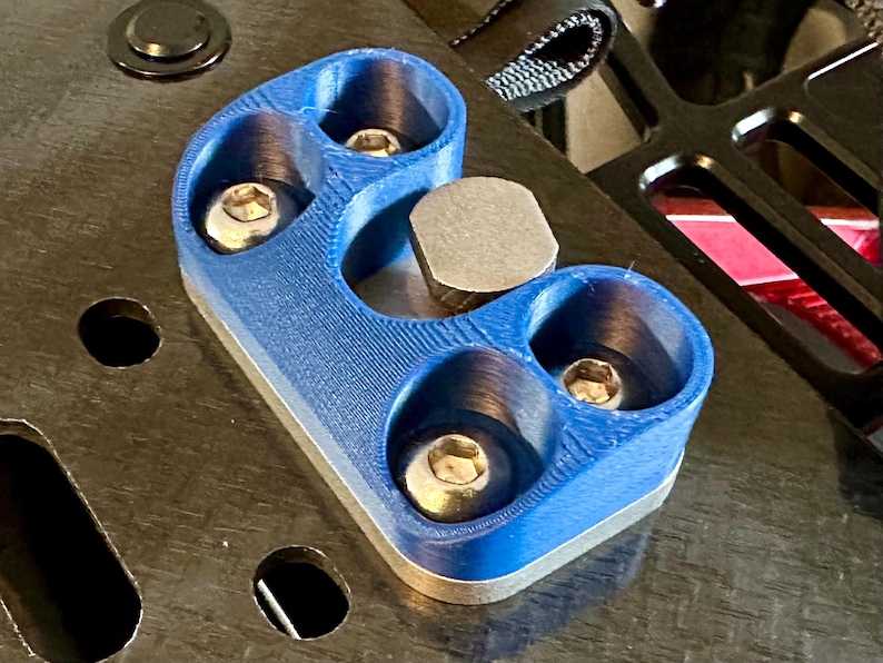 May include: A blue plastic part with four screw holes and a silver metal piece in the center. The part is mounted on a black surface.