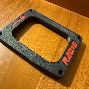 May include: A black rectangular frame with rounded corners. The frame has an arrow pointing right and the word "RAD3D" in red. There are four holes in the corners. The frame is on a wooden surface.
