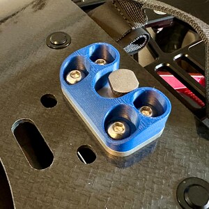 May include: A blue plastic mount with four silver screws and a silver metal plate. The mount is attached to a black surface with a woven pattern.
