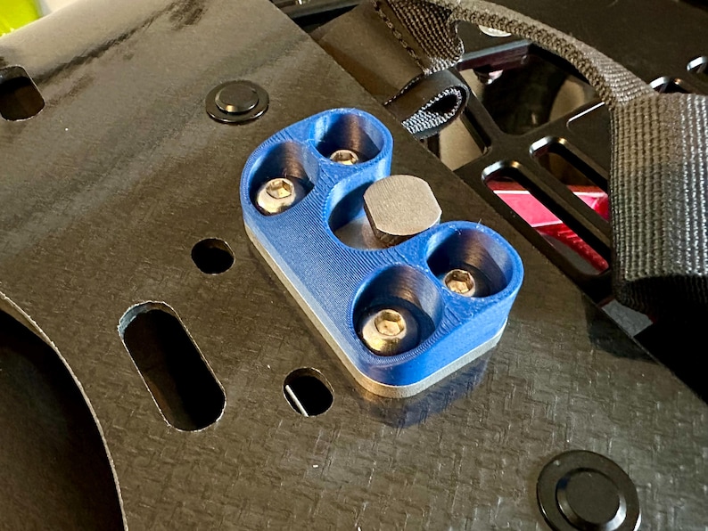 May include: A blue plastic mount with four silver screws and a silver metal plate. The mount is attached to a black surface with a woven pattern.
