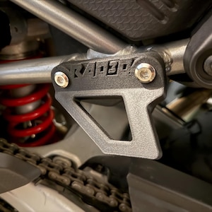 以下が含まれることがあります： 「RAPID」の文字がプリントされた黒いオートバイ部品のクローズアップ。 部品は2本の銀色のボルトで銀色のフレームに取り付けられています。 赤いスプリングとチェーンが背景に見えます。