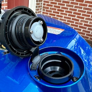 May include: Close-up of a blue gas tank with a black gas cap open. The gas cap has a white plastic vent and is secured with screws.
