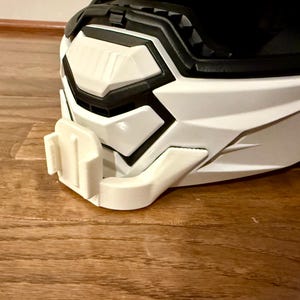 May include: Close-up of a white and black motorcycle helmet with a camera mount attached. The helmet has a geometric design with sharp angles and a visor. The camera mount is white and designed to hold a small action camera.