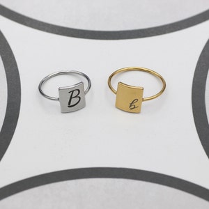 Può includere: Due anelli sottili, uno argentato e uno dorato, ciascuno con un ciondolo quadrato con un'iniziale. L'anello argentato mostra la lettera "B", mentre quello dorato ha una "b" in corsivo. Entrambi gli anelli sono su sfondo bianco e grigio.