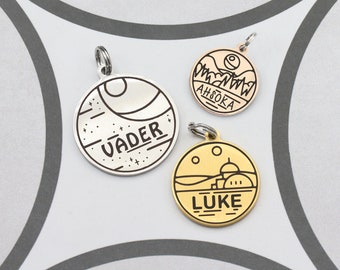May include: Three round metal pet tags with black engraved designs. The first tag is silver and features a Death Star design with the name "VADER". The second tag is rose gold and features a mountain range design with the name "AHsoka". The third tag is gold and features a desert landscape with the name "LUKE".
