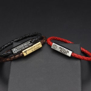 Könnte beinhalten: Zwei Lederarmbänder mit gravierten Metallplatten. Ein Armband ist aus schwarzem Leder mit einer goldenen Platte, auf der "COLIN MILLER ICE 123.456.7890 Krankenkarte im Portemonnaie" steht. Das andere Armband ist aus rotem Leder mit einer silbernen Platte, auf der "MEDIZINISCHE ALARM" steht.