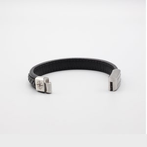 May include: Black leather bracelet with a silver compass charm and a silver clasp. The bracelet is braided and has a silver inscription on the clasp.