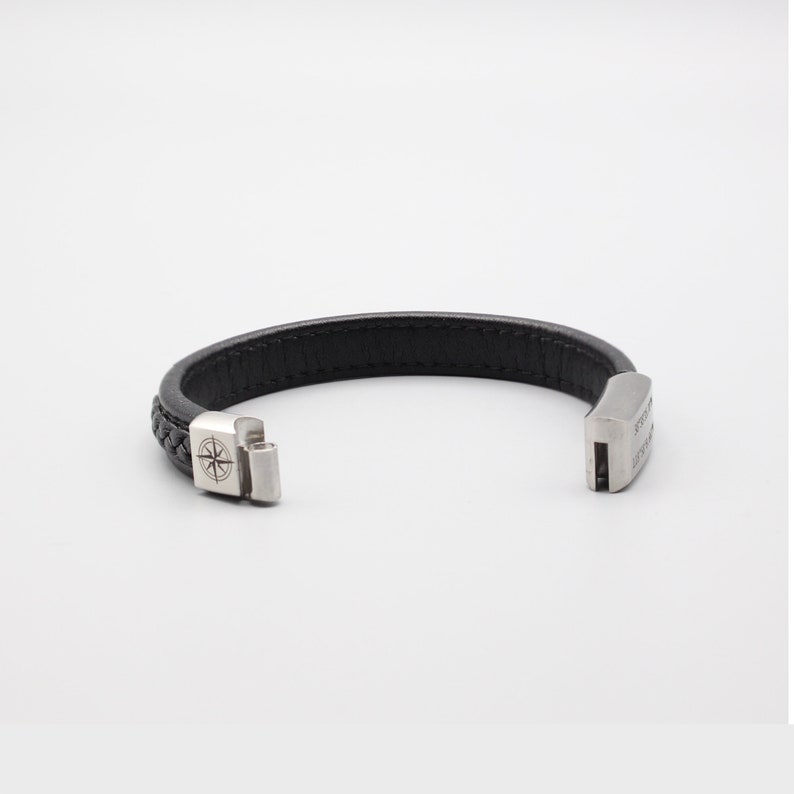 May include: Black leather bracelet with a silver compass charm and a silver clasp. The bracelet is braided and has a silver inscription on the clasp.