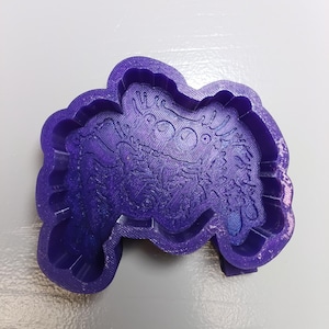 May include: A purple cookie cutter with a textured finish, featuring an intricate embossed design of a spider with multiple eyes and detailed legs. The tool has a raised outer wall for cutting and an inner stamp for creating impressions in dough.