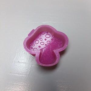 May include: A bright pink, glittery mushroom-shaped cookie cutter with a raised rim and a detailed interior design featuring a cap with circular spots and a central stem. The object has a textured, 3D-printed finish with visible layer lines.