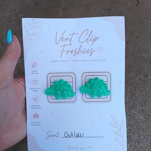 May include: A card with two green dinosaur-shaped vent clip air fresheners. The card reads "Vent Clip Freshies" and "Scent: Outlaw". The card also has text that says "Good Scents, Good Vibes, Good Days".