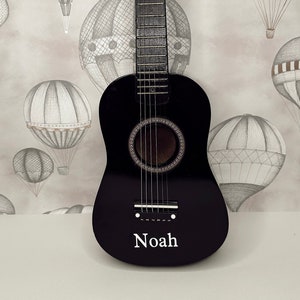 May include: A black acoustic guitar with a white name "Noah" on the front. The guitar is in front of a light gray background with a pattern of black and white hot air balloons.