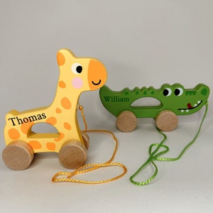 Op de afbeelding: Twee houten trek speelgoedjes, een gele giraffe en een groene krokodil, met touwtjes eraan vastgemaakt. De giraffe heeft de naam "Thomas" erop staan en de krokodil heeft de naam "William" erop staan.