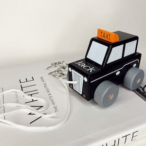 May include: Black wooden toy car with a white 'Jack' name and a yellow 'Taxi' sign on top. The car has gray wheels and a white string attached to the front.