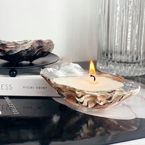 May include: A white candle burning inside a natural oyster shell with a silver finish. The shell is sitting on a black surface with a book and a clear glass bottle in the background.