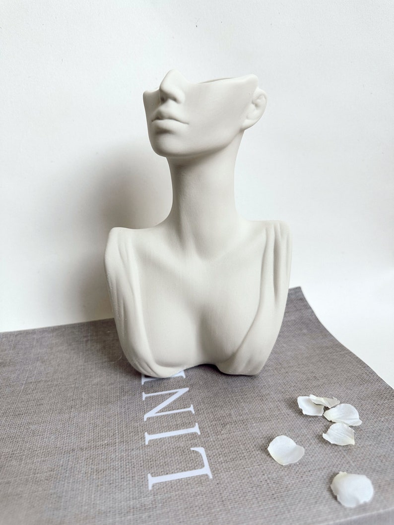 May include: White ceramic planter shaped like a woman's bust with a hollowed-out face. The planter is sitting on a gray textured surface with the word "LINE" printed in white.