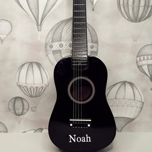 May include: A black acoustic guitar with a white name tag that says "Noah" on it. The guitar is in front of a background of hot air balloons.