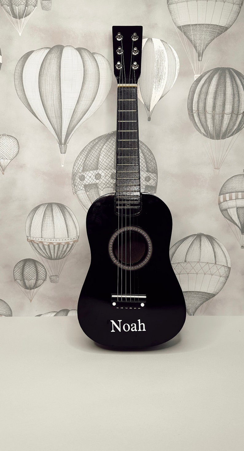 May include: A black acoustic guitar with a white name tag that says "Noah" on it. The guitar is in front of a background of hot air balloons.