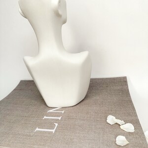 May include: White ceramic bust sculpture with a geometric design. The bust is sitting on a brown book with the word "LINEN" printed on the cover.